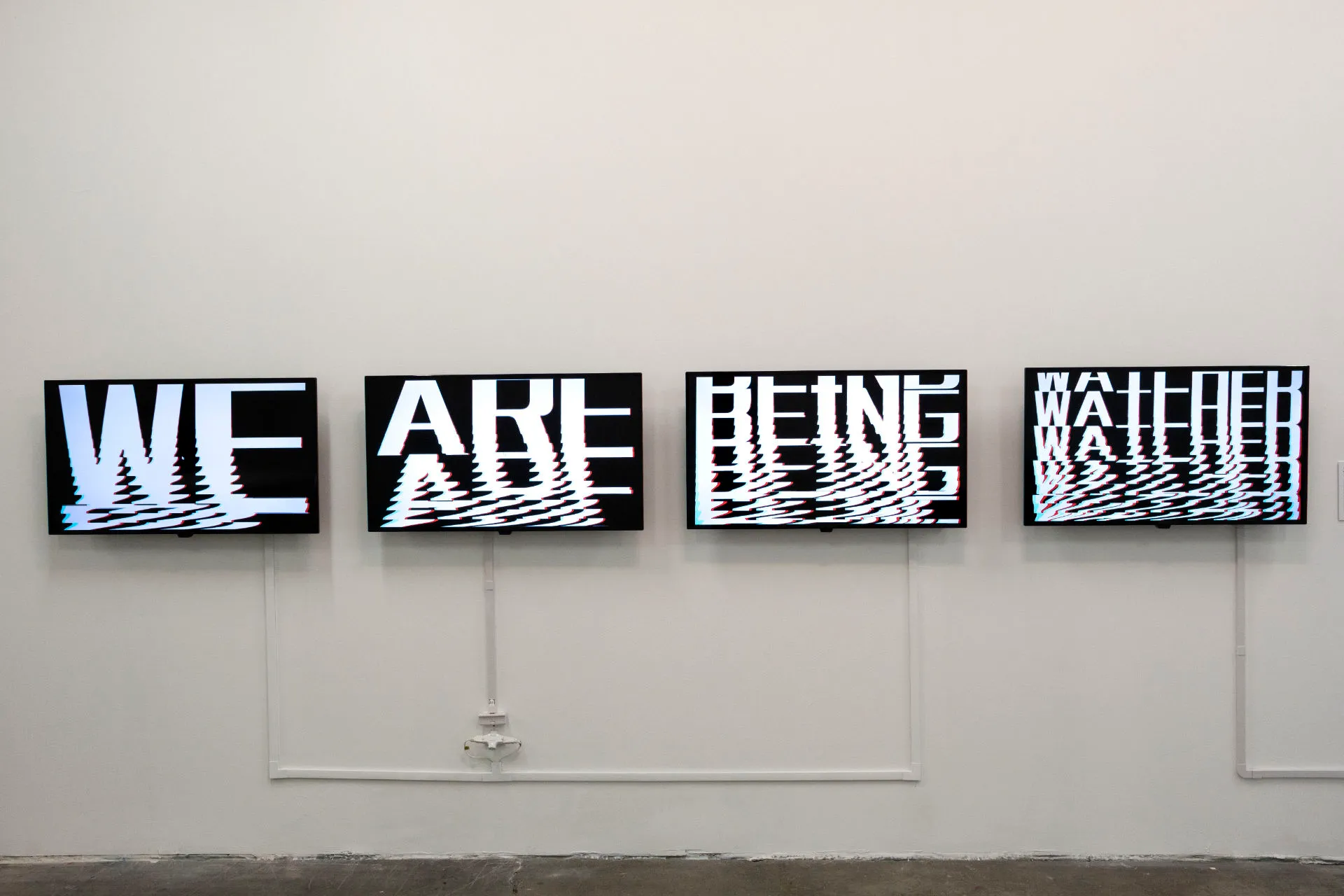 Four screens on a white wall that form the sentence WE ARE BEING WATCHED