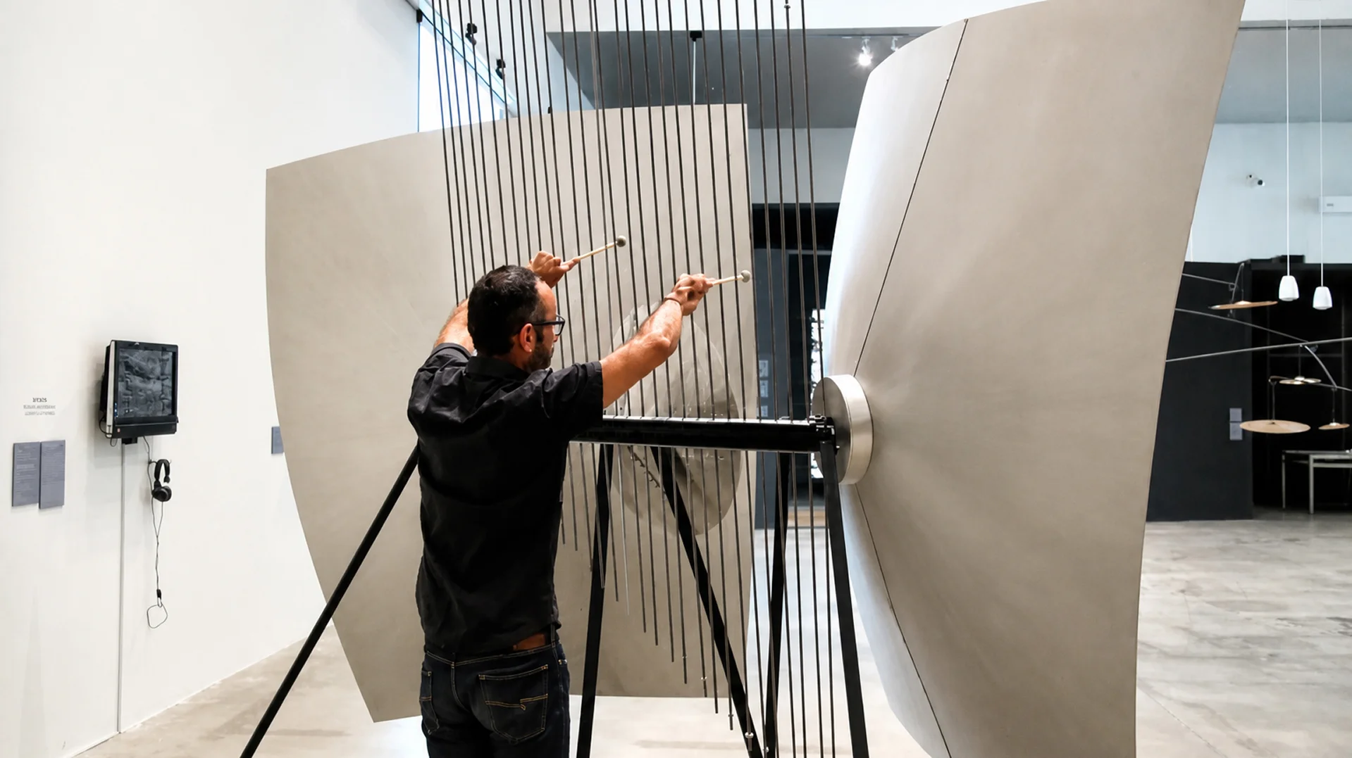 Man playing a large installation instrument with metal rods and wooden mallets in a modern gallery.
