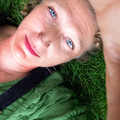 Woman with blue eyes and red lipstick lying on green grass wearing a green shirt and black strap.